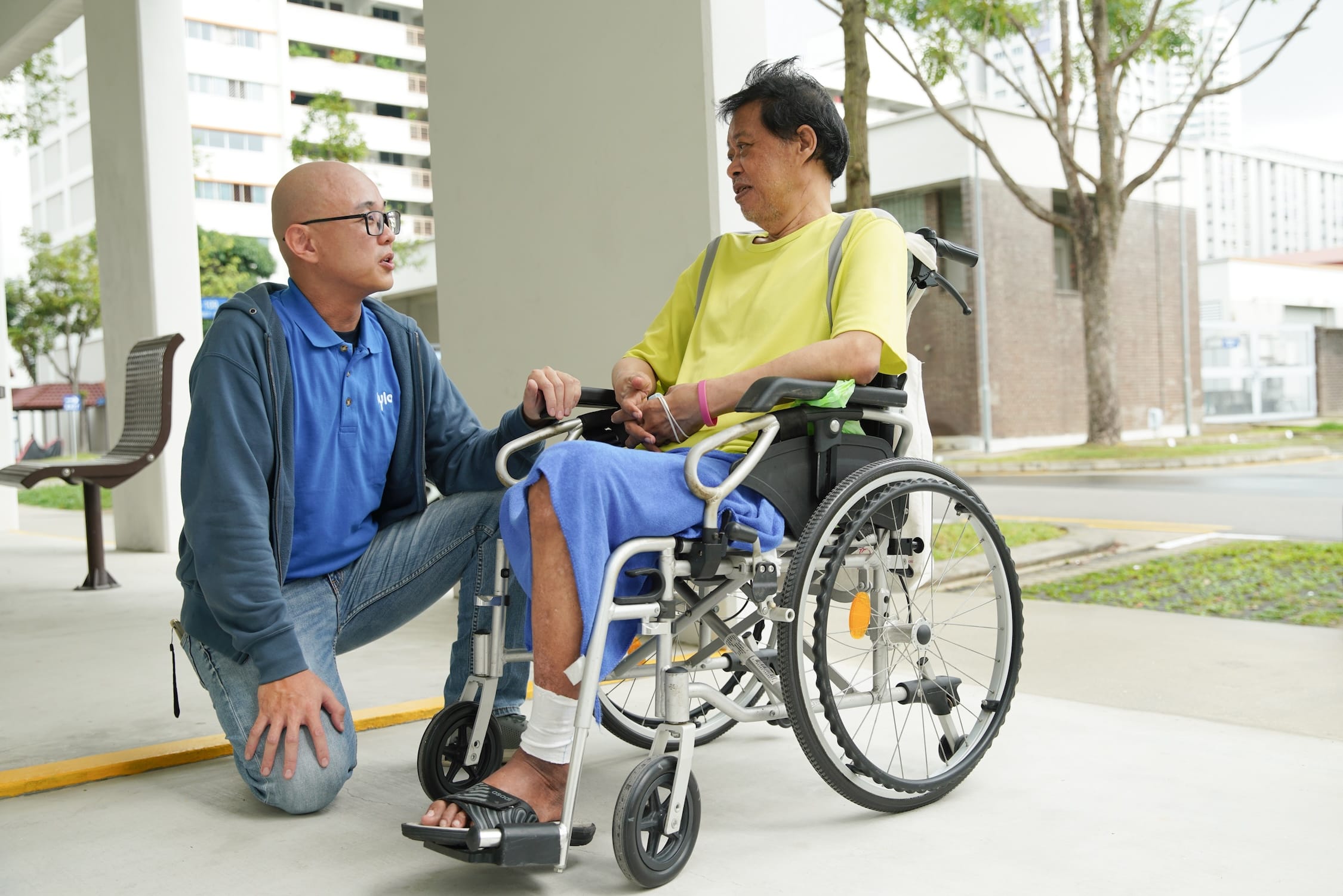 Bald man kneels, talking to man in wheelchair with bandaged leg, yellow shirt, blue blanket.